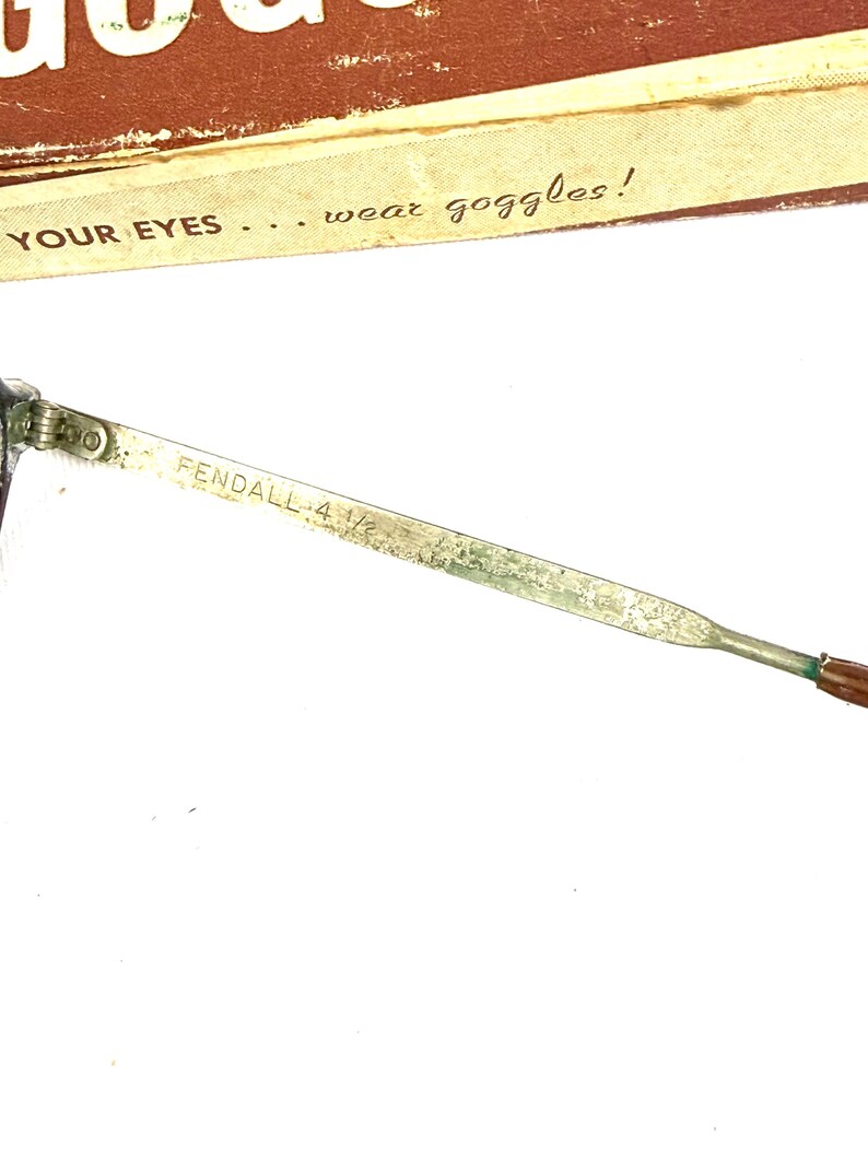 Pode incluir: Close-up de uma haste de &oacute;culos de seguran&ccedil;a vintage. A haste de metal est&aacute; marcada com "FENDALL 4 1/2". O texto "YOUR EYES... wear goggles!" &eacute; vis&iacute;vel em um fundo marrom.
