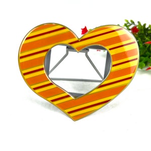 May include: A heart-shaped picture frame with a striped design in shades of orange and yellow. The frame is made of metal and has a stand on the back.