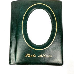 Peut inclure: Un album photo vert fonc&eacute; avec un cadre ovale dor&eacute; et l'inscription "Photo Album" en lettres dor&eacute;es. L'album a une texture lisse semblable &agrave; du cuir et un design classique.