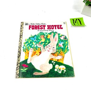 Puede incluir: Un pequeño libro dorado titulado "Forest Hotel: Una historia de conteo" con un conejo blanco de orejas largas y nariz rosada, rodeado de pájaros coloridos y hongos en un entorno de bosque verde.