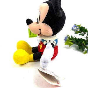 May include: A plush Mickey Mouse toy, featuring black ears, a tan face, and a red and white outfit. The toy has yellow feet and white gloves. A green tag is attached to the toy. The toy is sitting on a white surface.