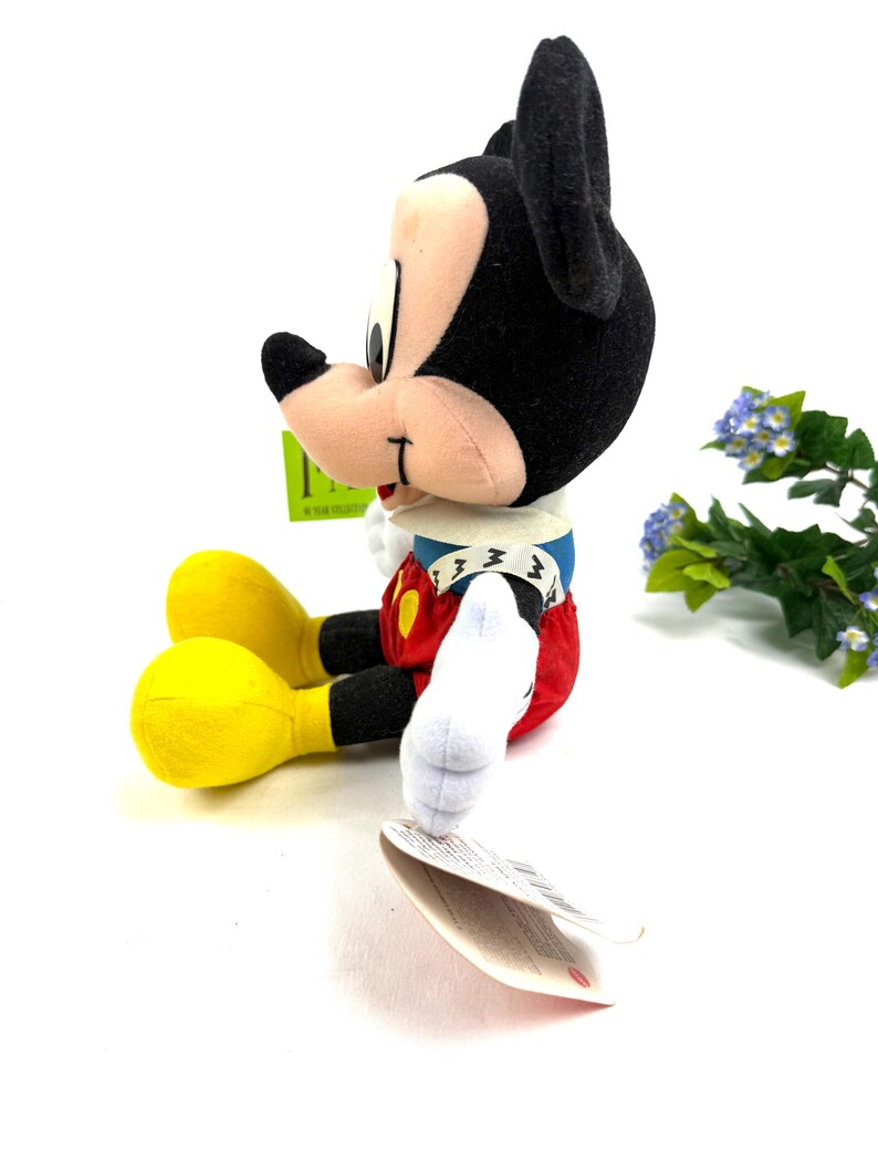 May include: A plush Mickey Mouse toy, featuring black ears, a tan face, and a red and white outfit. The toy has yellow feet and white gloves. A green tag is attached to the toy. The toy is sitting on a white surface.