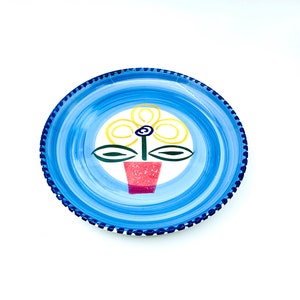 May include: A blue and white ceramic plate with a stylized flower design in yellow, green, and red. The plate has a blue rim with a white and blue dotted pattern.