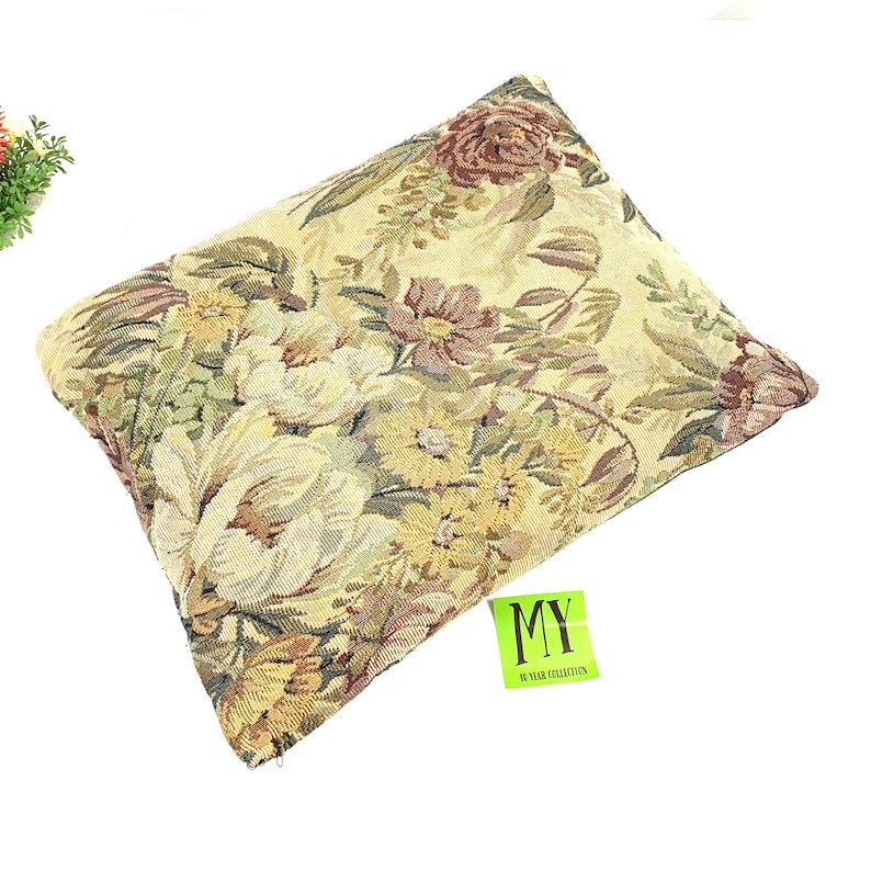 May include: A rectangular pillow with a floral tapestry design in shades of green, brown, and yellow. The pillow has a zipper closure and a tag that reads "MY 70 YEAR COLLECTION".