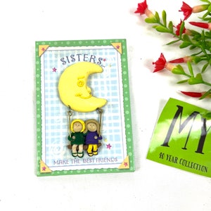 May include: A yellow crescent moon with two girls holding onto ropes hanging from the moon. The girls are wearing purple and green dresses. The card reads "Sisters... Make the best friends".