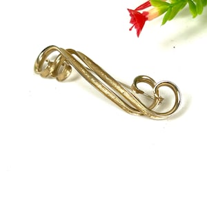 May include: A gold-toned brooch with a swirling, abstract design. The brooch is made of two pieces of metal that are intertwined.