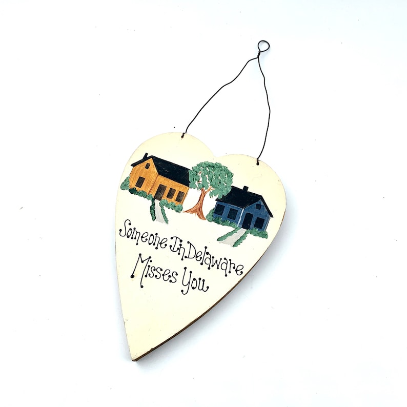 May include: A white heart-shaped wooden sign with a painted scene of two houses and a tree. The text "Someone in Delaware Misses You" is written in black cursive.