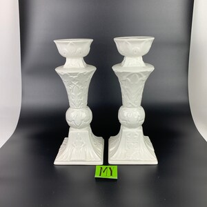May include: A pair of white ceramic candle holders with an ornate design. The holders have a square base and a tall, slender body with a decorative pattern.
