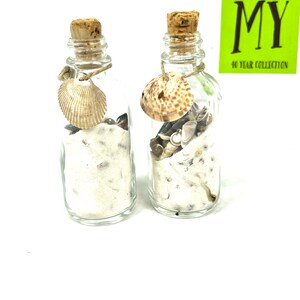 May include: Two small glass bottles filled with sand and seashells. The bottles have cork stoppers and are tied with twine. Each bottle has a seashell tied to the twine.