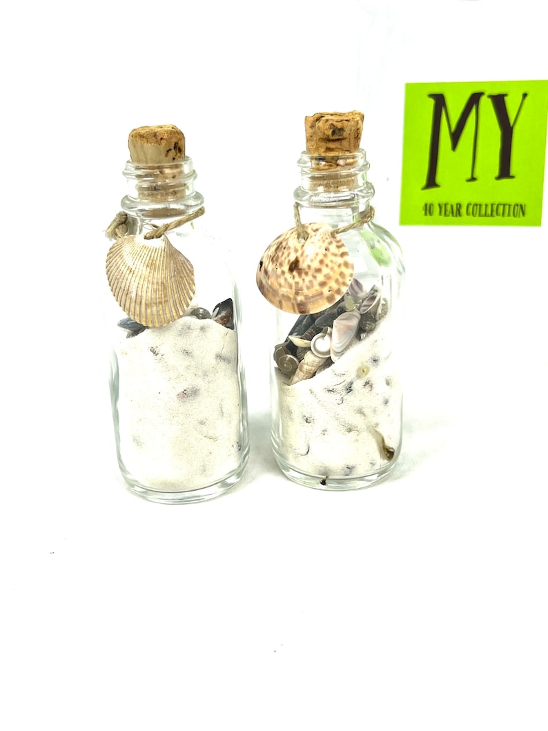 May include: Two small glass bottles filled with sand and seashells. The bottles have cork stoppers and are tied with twine. Each bottle has a seashell tied to the twine.