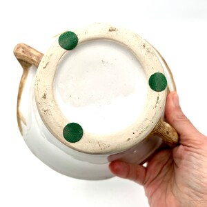 May include: A white ceramic pitcher with a brown handle and spout. The bottom of the pitcher is visible, showing a white interior and three green felt pads. The pitcher is held by a person's hand.