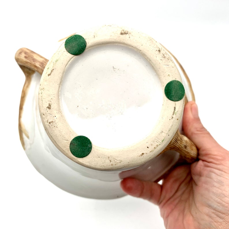 May include: A white ceramic pitcher with a brown handle and spout. The bottom of the pitcher is visible, showing a white interior and three green felt pads. The pitcher is held by a person's hand.