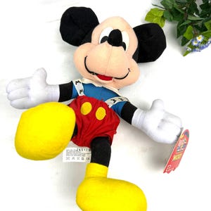 May include: A Mickey Mouse plush toy with black ears, a pink face, and white gloves. Mickey wears a blue jacket, red shorts with yellow buttons, and yellow shoes. The toy has a tag that reads "Mickey". A green square tag reads "MY 90 YEAR COLLECTION".