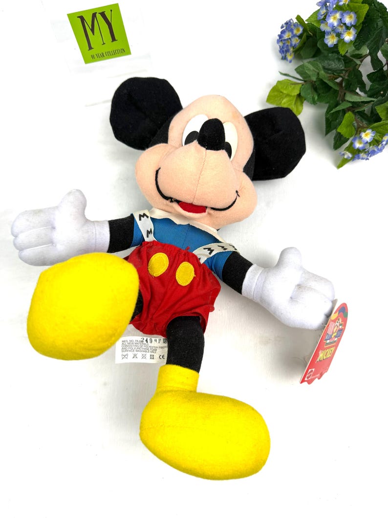 May include: A Mickey Mouse plush toy with black ears, a pink face, and white gloves. Mickey wears a blue jacket, red shorts with yellow buttons, and yellow shoes. The toy has a tag that reads "Mickey". A green square tag reads "MY 90 YEAR COLLECTION".