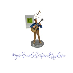 May include: A figurine of a man playing a guitar, dressed in a blue shirt and tan trousers. The figurine stands on a silver base with the words "Love Me Tender". A tag with "MY" and "Vintage Collection" is attached. The Etsy shop name "My40YearCollection.Etsy.com" is written in cursive.