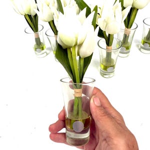 Peut inclure: Plusieurs bouquets de tulipes blanches dans des verres &agrave; shot transparents. Chaque tulipe a des tiges et des feuilles vertes. Les verres &agrave; shot contiennent un liquide jaune et sont attach&eacute;s avec de la ficelle. Une main tient un verre.