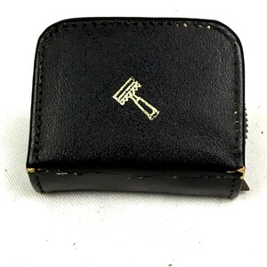 May include: A black, rectangular case with a gold-colored razor emblem. The case has a zipper closure and appears to be made of leather. The razor emblem is a stylized depiction of a razor.