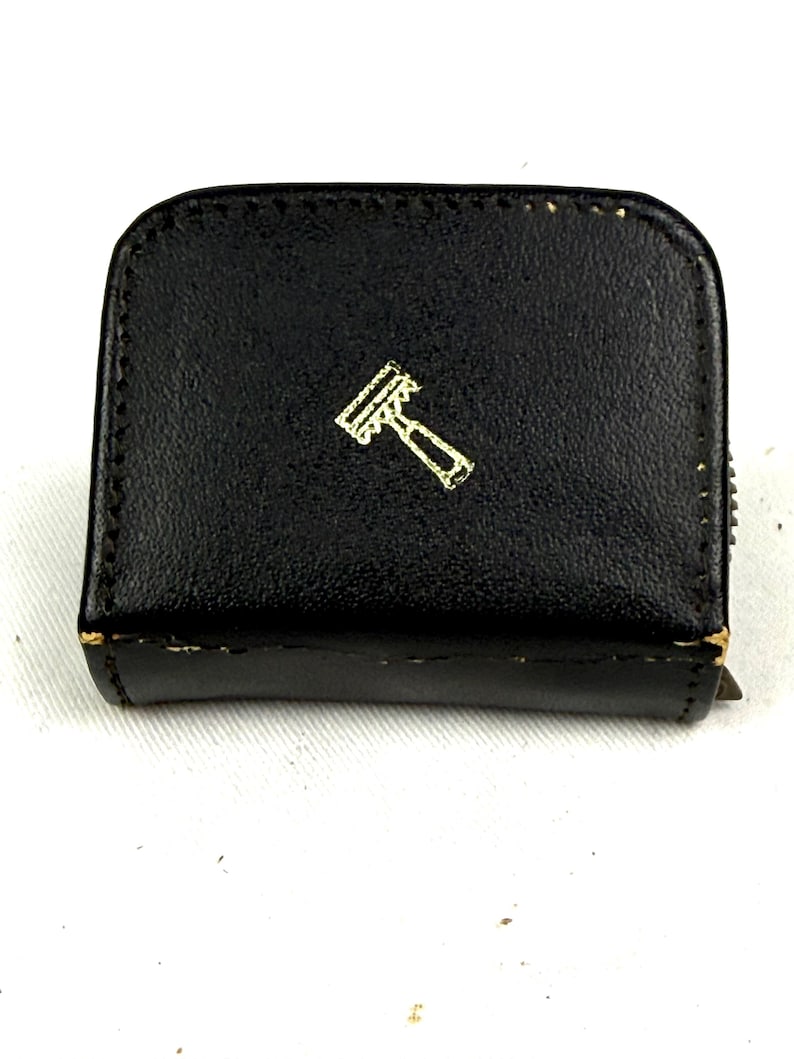 May include: A black, rectangular case with a gold-colored razor emblem. The case has a zipper closure and appears to be made of leather. The razor emblem is a stylized depiction of a razor.