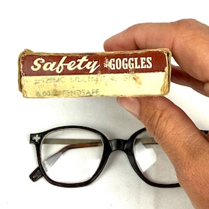 Pode incluir: &Oacute;culos vintage com arma&ccedil;&atilde;o preta e lentes transparentes. Uma pequena caixa de cart&atilde;o com a etiqueta "Safety Goggles" tamb&eacute;m est&aacute; presente. A caixa tem texto desbotado e um esquema de cores castanho e creme.