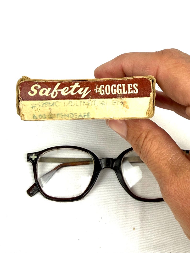 Pode incluir: &Oacute;culos vintage com arma&ccedil;&atilde;o preta e lentes transparentes. Uma pequena caixa de cart&atilde;o com a etiqueta "Safety Goggles" tamb&eacute;m est&aacute; presente. A caixa tem texto desbotado e um esquema de cores castanho e creme.