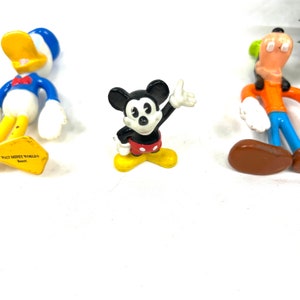 Może przedstawiać: Trzy plastikowe figurki postaci z Disneya: Myszka Miki w czerwonych spodenkach i ż&oacute;łtych butach, Kaczor Donald w niebieskiej marynarskiej koszuli i ż&oacute;łtych butach oraz Goofy w pomarańczowych spodniach i brązowych butach. Figurka Myszki Miki ma napis "Walt Disney World Resort" na spodzie.