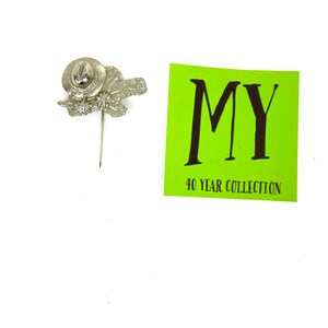 May include: Silver pin with a hat and leaves design. The pin has a small, round, silver base with a pin back. The pin is on a white background. The text "MY 40 YEAR COLLECTION" is on a green background.