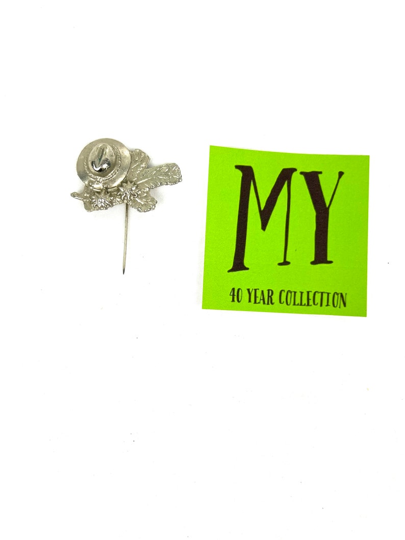 May include: Silver pin with a hat and leaves design. The pin has a small, round, silver base with a pin back. The pin is on a white background. The text "MY 40 YEAR COLLECTION" is on a green background.