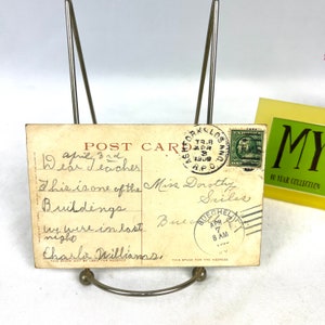 May include: Aged postcard with handwritten text and postmarks, displayed in a metal stand. The postcard features a green postage stamp and a postmark from Los Angeles, California, dated April 8, 1909. The text includes "Dear Teacher".