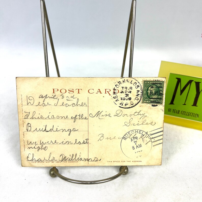 May include: Aged postcard with handwritten text and postmarks, displayed in a metal stand. The postcard features a green postage stamp and a postmark from Los Angeles, California, dated April 8, 1909. The text includes "Dear Teacher".