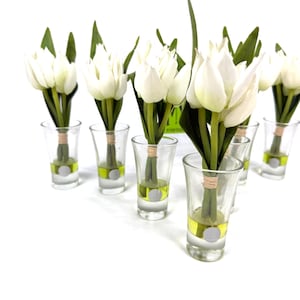 Peut inclure: Plusieurs bouquets de tulipes blanches dans des verres &agrave; shot transparents. Chaque verre contient un peu de liquide jaune et un autocollant argent&eacute;. Les tiges des tulipes sont li&eacute;es avec de la ficelle, et des feuilles vertes sont visibles.