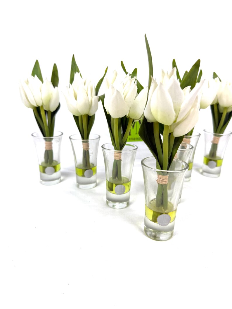 Peut inclure: Plusieurs bouquets de tulipes blanches dans des verres &agrave; shot transparents. Chaque verre contient un peu de liquide jaune et un autocollant argent&eacute;. Les tiges des tulipes sont li&eacute;es avec de la ficelle, et des feuilles vertes sont visibles.
