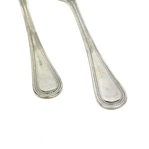 May include: Two shiny, silver spoons with a classic design. The spoon bowls are rounded, and the handles have a decorative beaded border. The spoons are set against a bright white background, highlighting their polished surfaces.