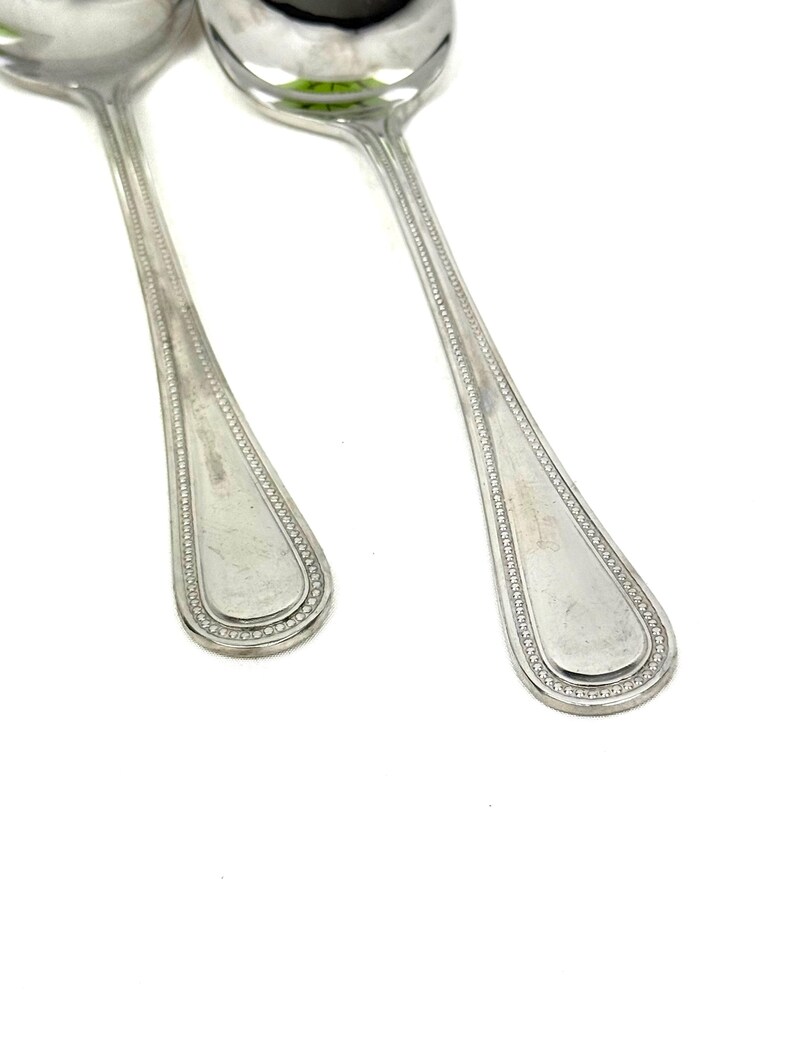 May include: Two shiny, silver spoons with a classic design. The spoon bowls are rounded, and the handles have a decorative beaded border. The spoons are set against a bright white background, highlighting their polished surfaces.