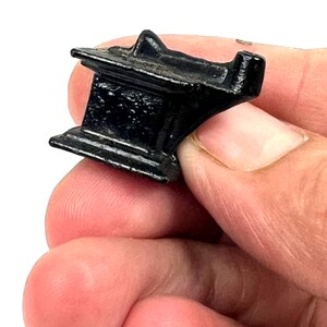 May include: A small, black metal anvil with a rounded top and a flat base. The anvil is held in a person's hand.