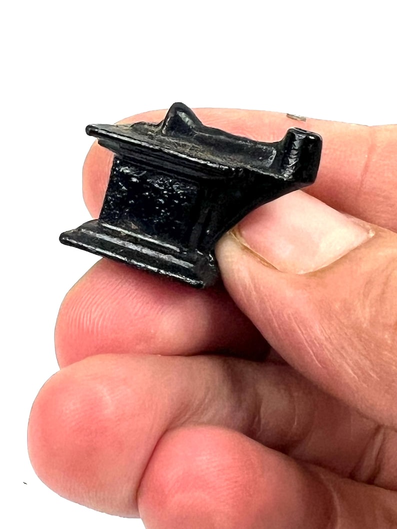 May include: A small, black metal anvil with a rounded top and a flat base. The anvil is held in a person's hand.