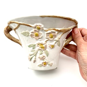 May include: White ceramic planter with embossed floral design. The planter features a light brown rim and handles, with delicate pink and yellow flowers and green leaves. The planter is held by a hand.