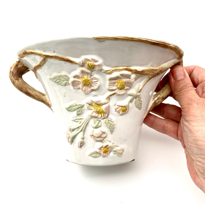 May include: White ceramic planter with embossed floral design. The planter features a light brown rim and handles, with delicate pink and yellow flowers and green leaves. The planter is held by a hand.
