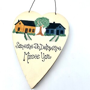 May include: A white heart-shaped wooden sign with a painted scene of two houses and a tree. The text "Someone in Delaware Misses You" is written in black paint.
