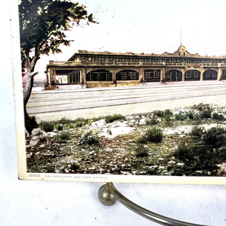 May include: A vintage postcard with a colourised image of a building with arched windows and a long facade. The text "THE ESCALANTE ASH FORK ARIZONA" is printed at the bottom. A tree is on the left. The postcard is displayed on a metal stand.
