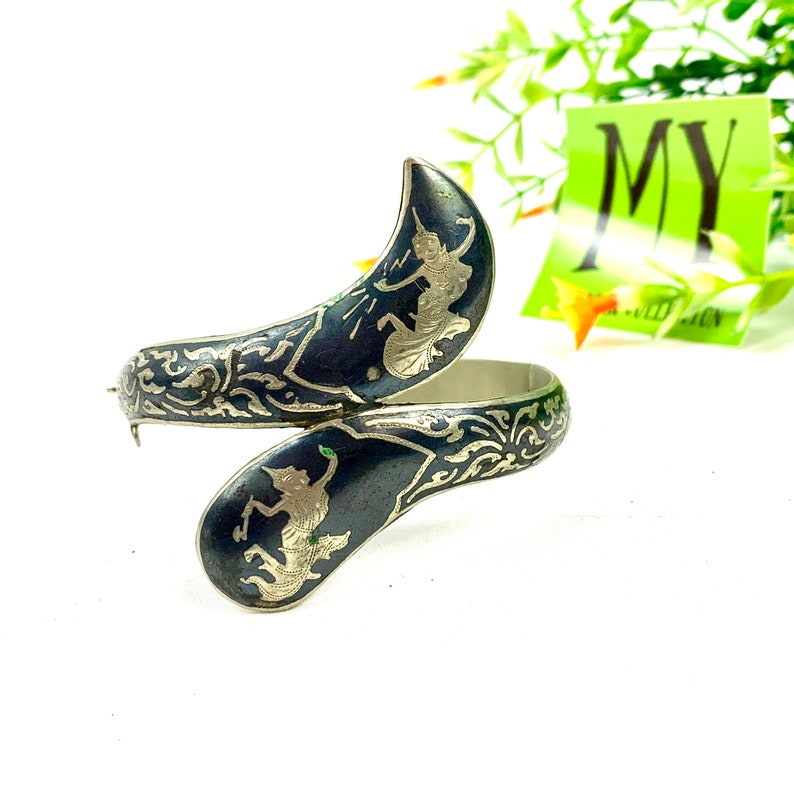 May include: A silver bangle bracelet with black enamel and intricate silver filigree designs. The bracelet features two figures of dancers in silver.