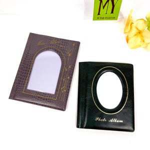 Peut inclure: Deux albums photo. L'un est marron fonc&eacute; avec une ouverture photo rectangulaire et des accents dor&eacute;s. L'autre est vert fonc&eacute; avec une ouverture photo ovale et les mots "Photo Album" en or. Les albums sont sur une surface blanche.