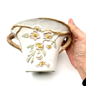 May include: White ceramic vase with embossed floral design. The vase features a light brown handle and rim, with delicate pink and yellow flowers and green leaves. The vase is held by a hand.