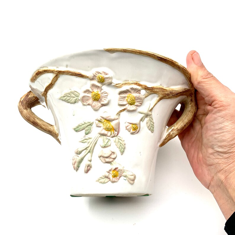 May include: White ceramic vase with embossed floral design. The vase features a light brown handle and rim, with delicate pink and yellow flowers and green leaves. The vase is held by a hand.