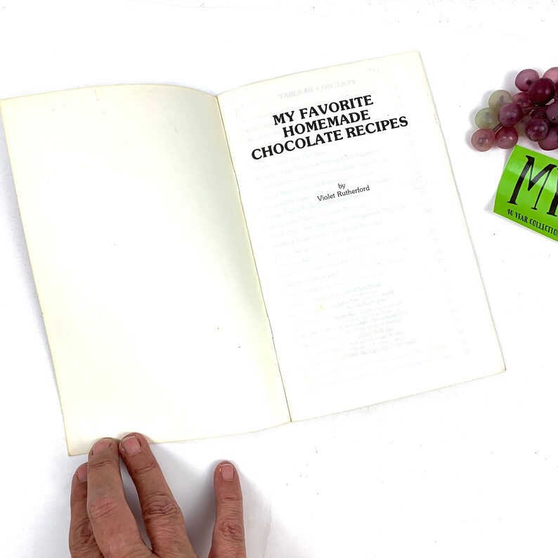 May include: An open book titled "My Favorite Homemade Chocolate Recipes" by Violet Rutherford. The book is open to the title page and is sitting on a white surface.