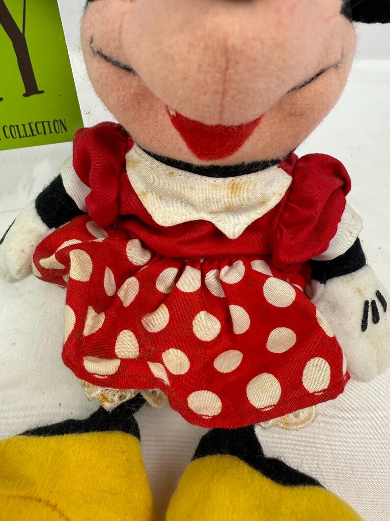 May include: A vintage Minnie Mouse plush doll. The doll features a red dress with white polka dots, a white collar, and black and white striped sleeves. The doll has yellow shoes and white gloves. The doll's face is pink with a red smile.