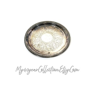 May include: A circular, silver-plated serving tray featuring an elaborate floral design in the centre. The tray has a detailed, rope-style edge and a slightly tarnished look. The text "My40yearCollection.Etsy.Com" is written in cursive at the bottom.