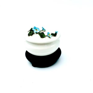 May include: A white ceramic trinket box with a floral design. The lid is decorated with blue forget-me-not flowers and green leaves. The box sits on a black wooden base.
