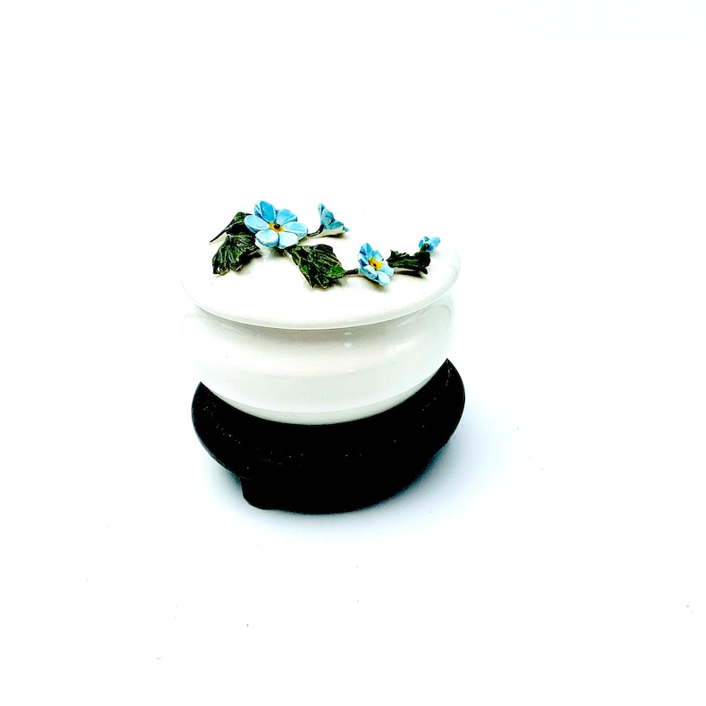 May include: A white ceramic trinket box with a floral design. The lid is decorated with blue forget-me-not flowers and green leaves. The box sits on a black wooden base.