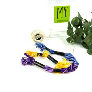 May include: Four spools of embroidery floss in purple, yellow, and blue colors. The spools are wrapped around black cardboard with white text that reads "DMC 100% Cotton Embroidery Floss". The spools are on a white surface with a white ball of yarn and a green plant in the background.