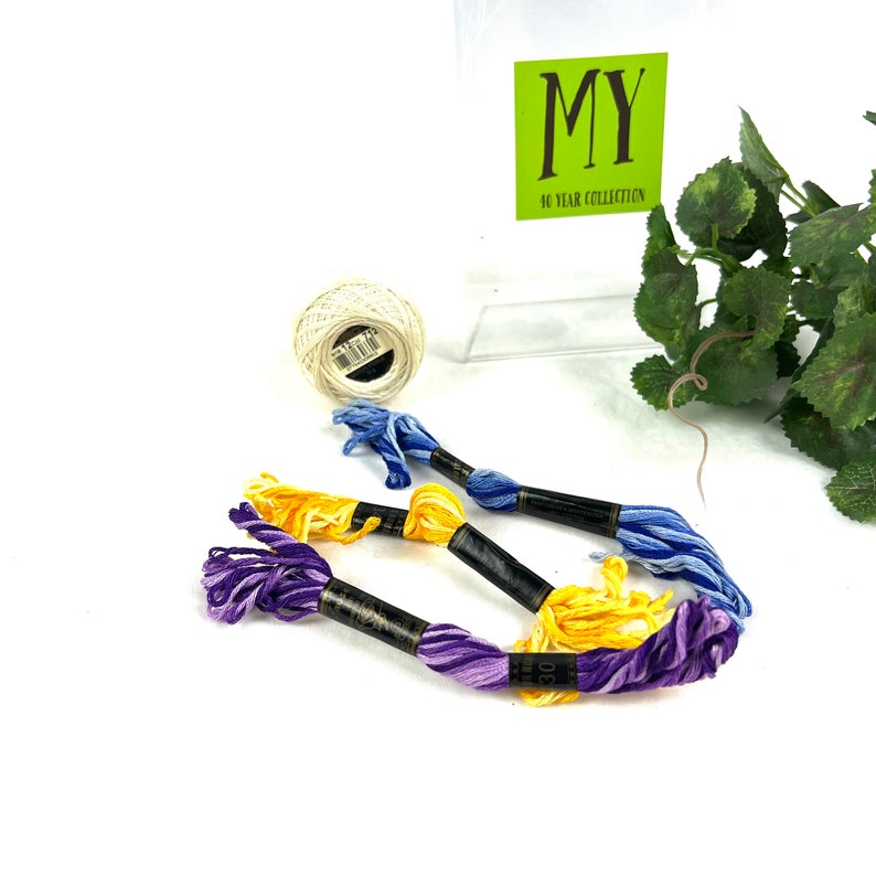 May include: Four spools of embroidery floss in purple, yellow, and blue colors. The spools are wrapped around black cardboard with white text that reads "DMC 100% Cotton Embroidery Floss". The spools are on a white surface with a white ball of yarn and a green plant in the background.